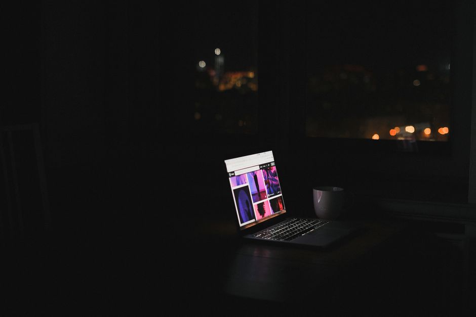 A laptop glows in a dark room at night with a cityscape through the window. Ideal for tech and solitude themes.