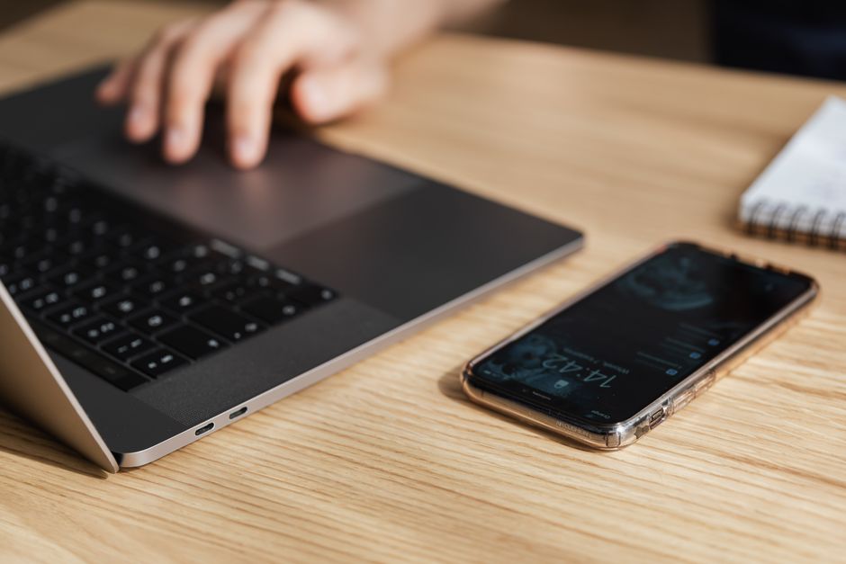 High angle of smartphone on wooden table near crop anonymous worker using touchpad on gray laptop